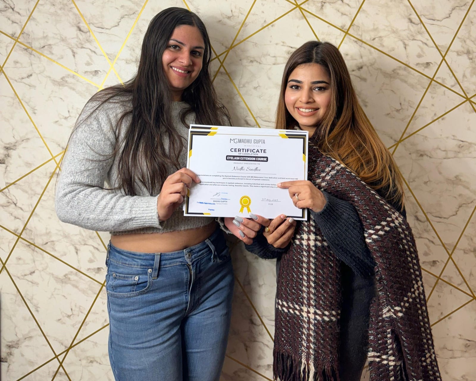 MG Makeovers Academy eyelash course graduates Two smiling MG Makeovers Academy graduates holding an Eyelash Extension Course certificate in front of a marble and gold wall