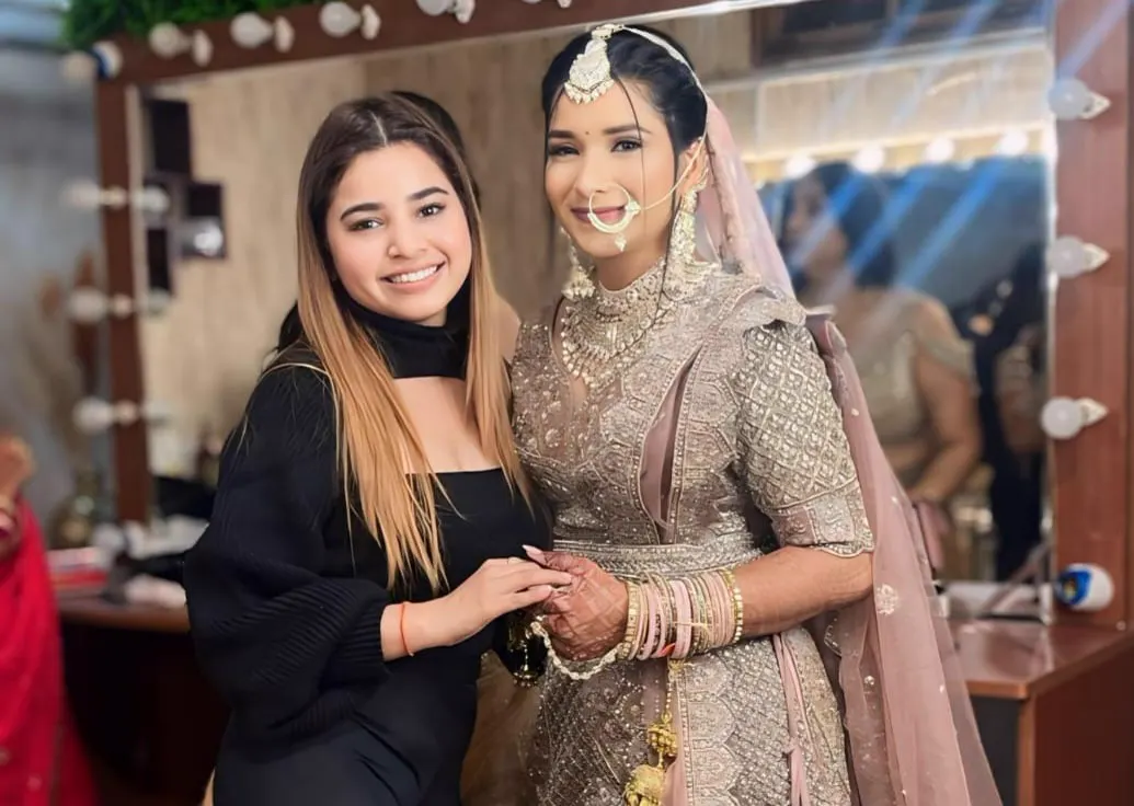 Madhu Gupta with her bride Madhu Gupta smiling beside a bride in a champagne-gold embroidered lehenga, pearl-drop nath and maang tikka in front of the MG Makeovers vanity mirrors