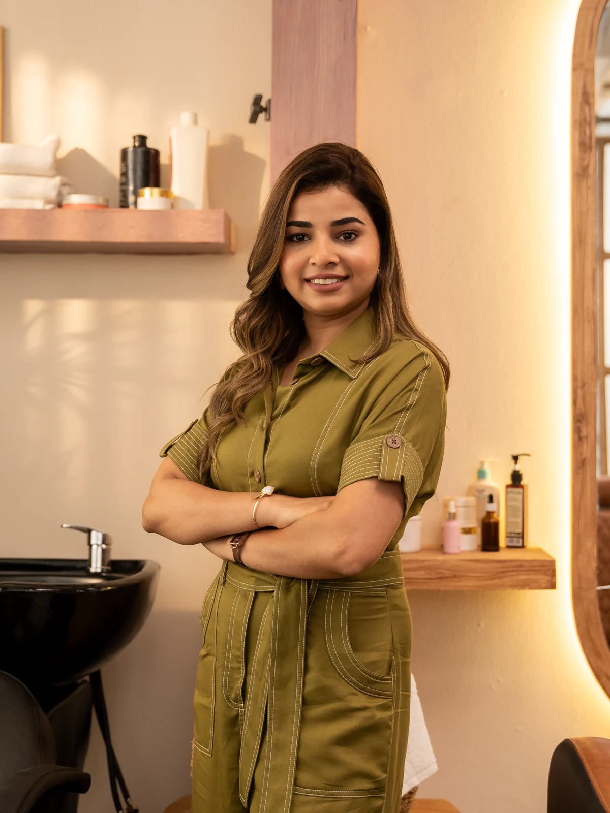 Madhu Gupta, founder of MG Makeovers Madhu Gupta, founder and lead artist of MG Makeovers, smiling with arms crossed in an olive utility jumpsuit inside her Gurugram studio