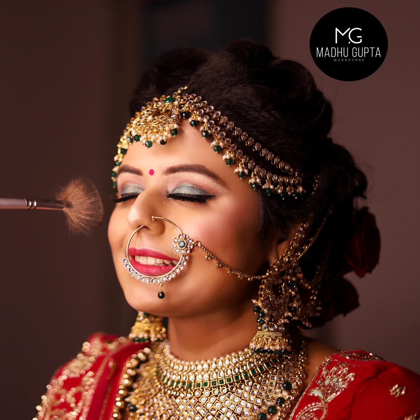 Bridal makeup touch-up Bride with eyes closed while a fan brush sweeps her cheek; gold matha patti with green beads, large nath, red lehenga and kundan necklace